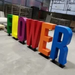 Multicolored large-format sheet metal letters spelling "Flower" in green, yellow, purple, red, orange, and blue